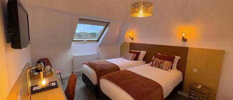 A hotel room with two beds, a desk with a chair, a television, a window, and a ceiling light fixture.