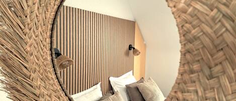 A circular mirror with a woven frame reflecting a room with a striped wall, a bed with pillows, and two wall-mounted lamps.