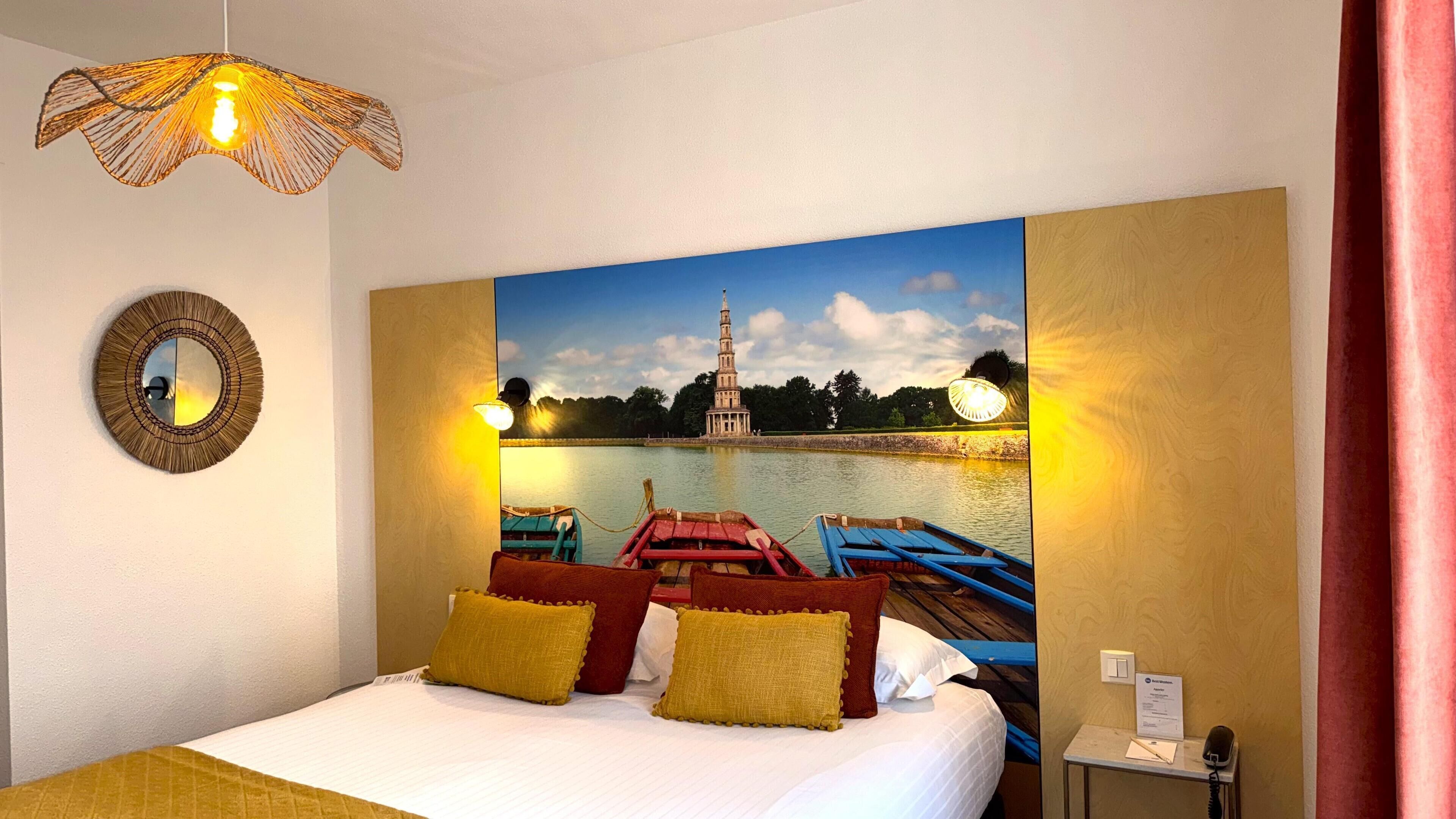 Habitación de hotel con una cama, un espejo redondo, una lámpara de pared, un cuadro de una torre junto a un lago y una mesita con un teléfono y un cartel.