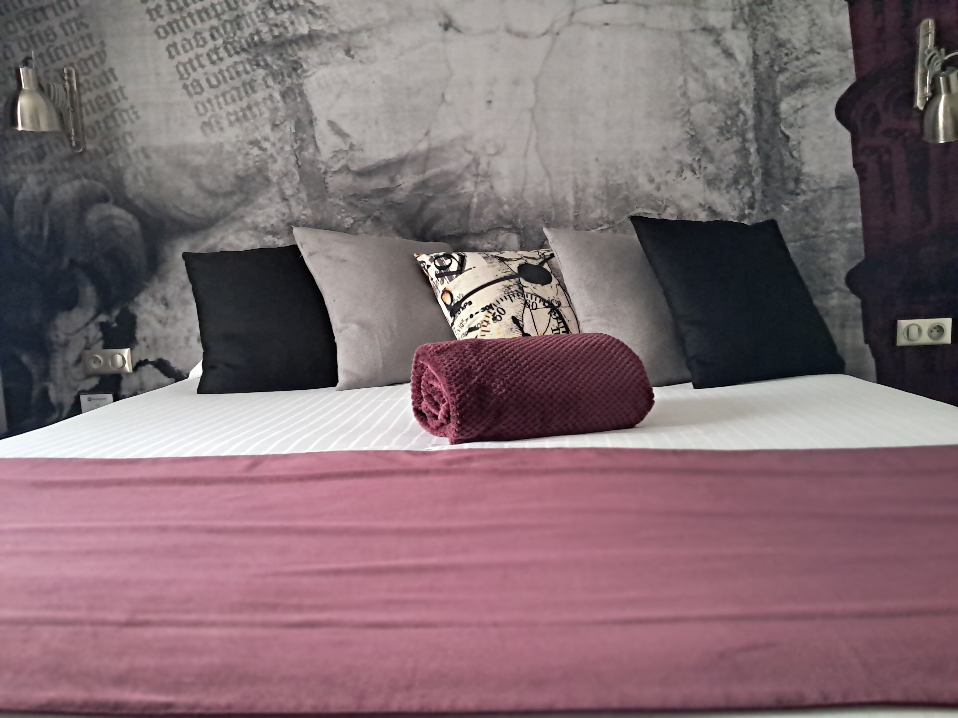Una cama con sábana blanca, una manta roja enrollada y tres almohadas en negro, gris y diseños estampados.