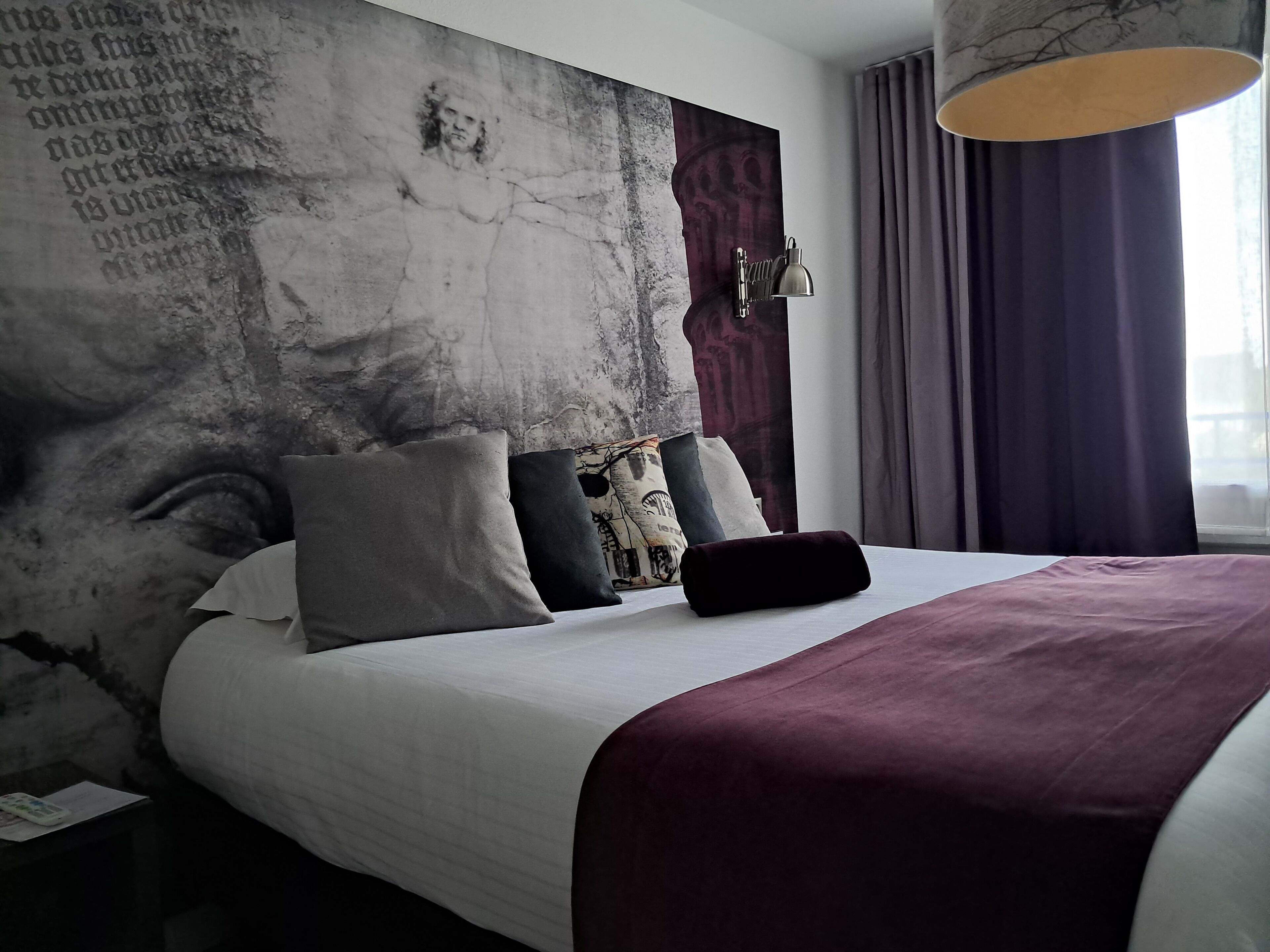 Una habitación de hotel moderna con una cama grande, varios almohadones, un cabecero con diseño texturizado, una mesita de noche y un ventanal con cortinas.
