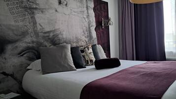 Una habitación de hotel moderna con una cama grande, varios almohadones, un cabecero con diseño texturizado, una mesita de noche y un ventanal con cortinas.