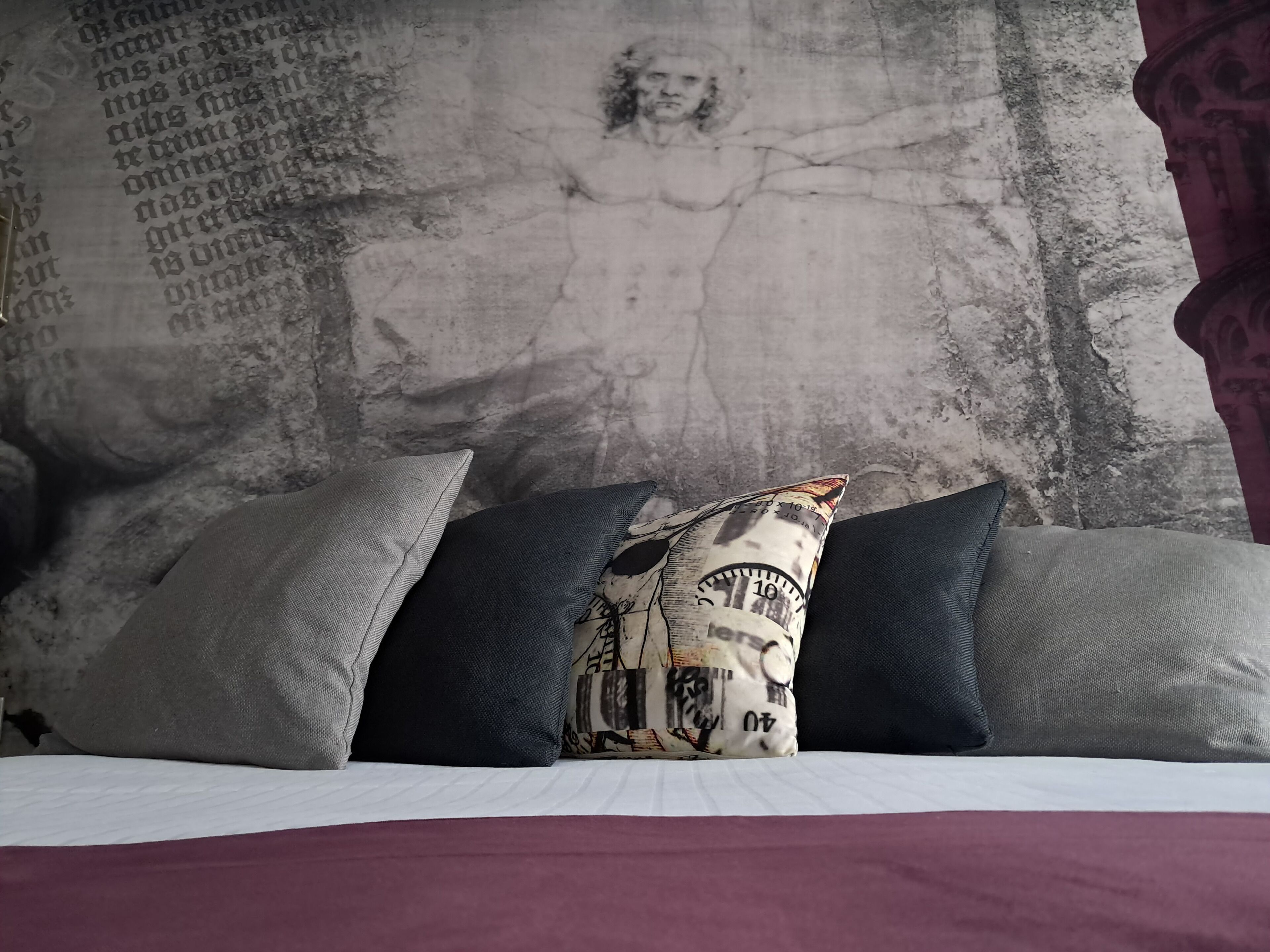 A bed with a white sheet and three pillows, one with a map design, against a textured wall with a mural.