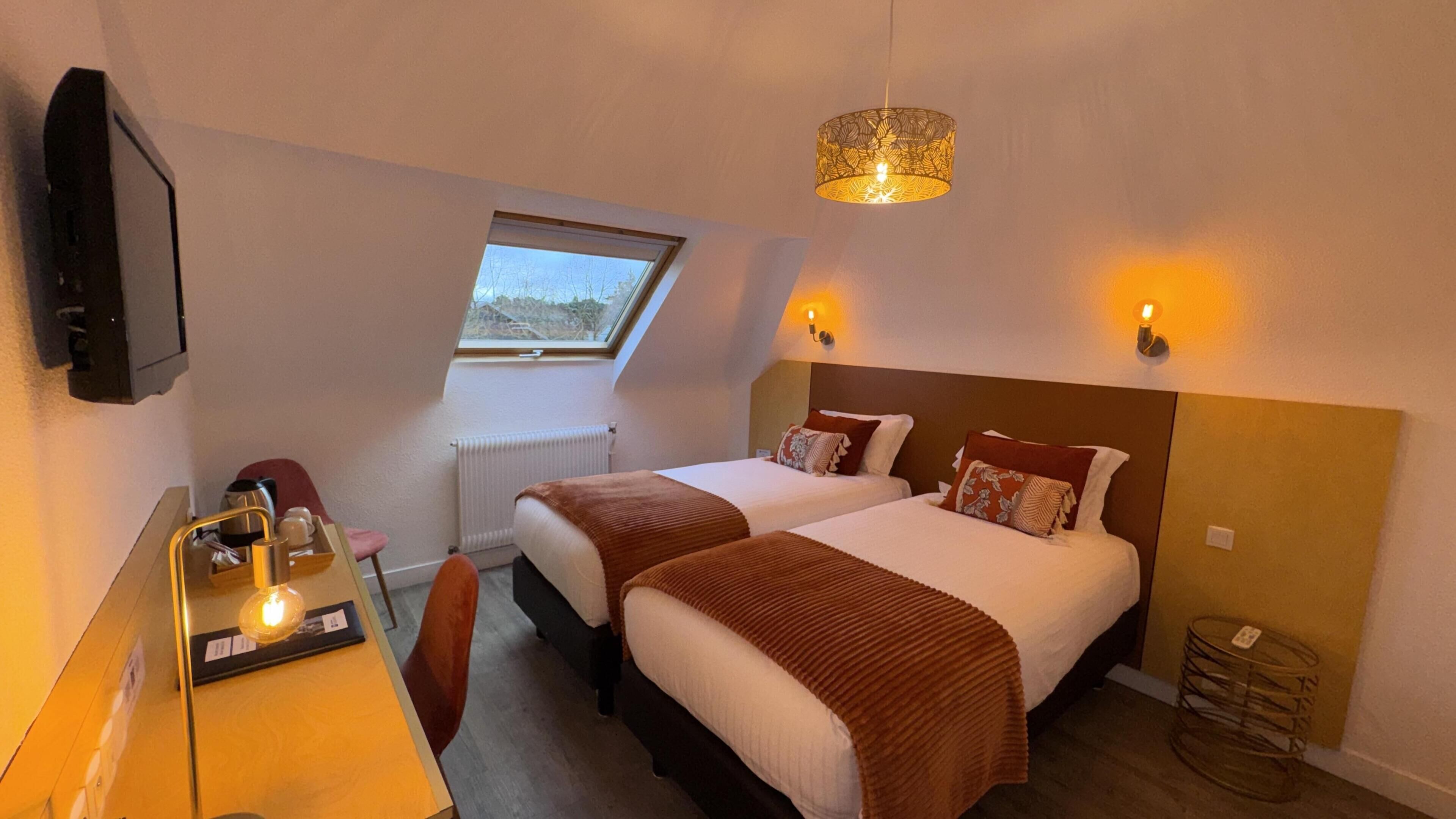 A hotel room with two beds, a desk with a chair, a television, a window, and a ceiling light fixture.