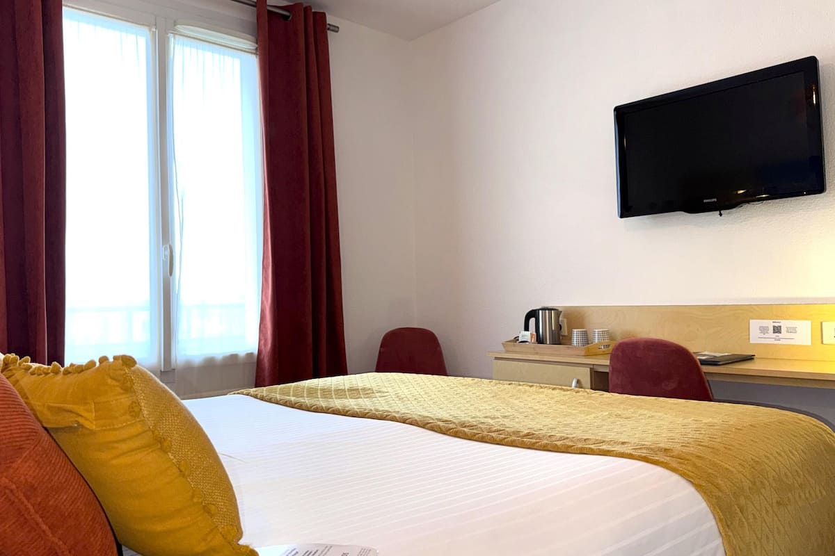 Une chambre d’hôtel comprenant un lit, un bureau avec un téléviseur à écran plat, une chaise et une fenêtre avec des rideaux.