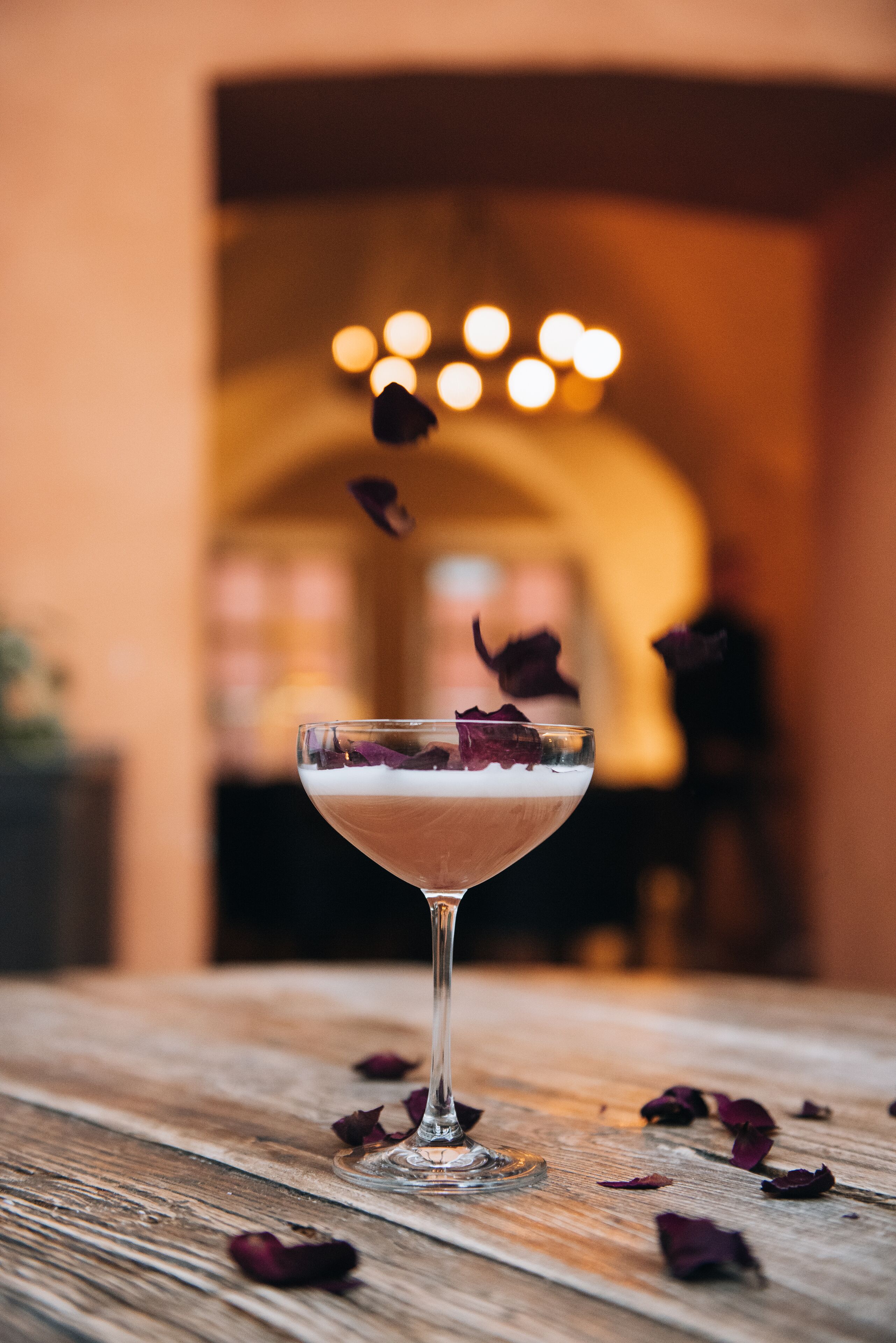 A cocktail glass with rose petals on a wooden surface.