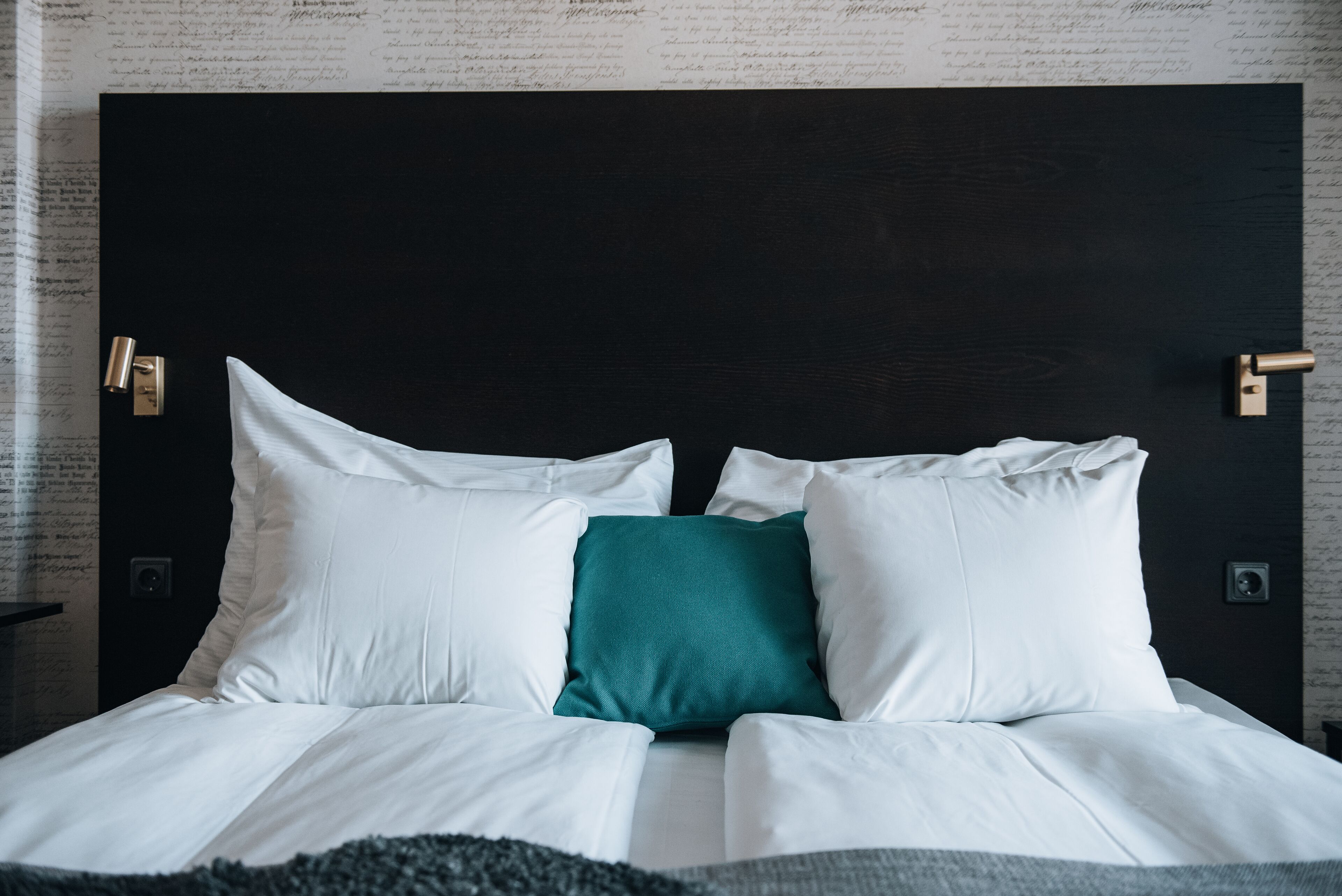 A bed with white and teal pillows, a dark headboard, and a wall with script-like patterns.