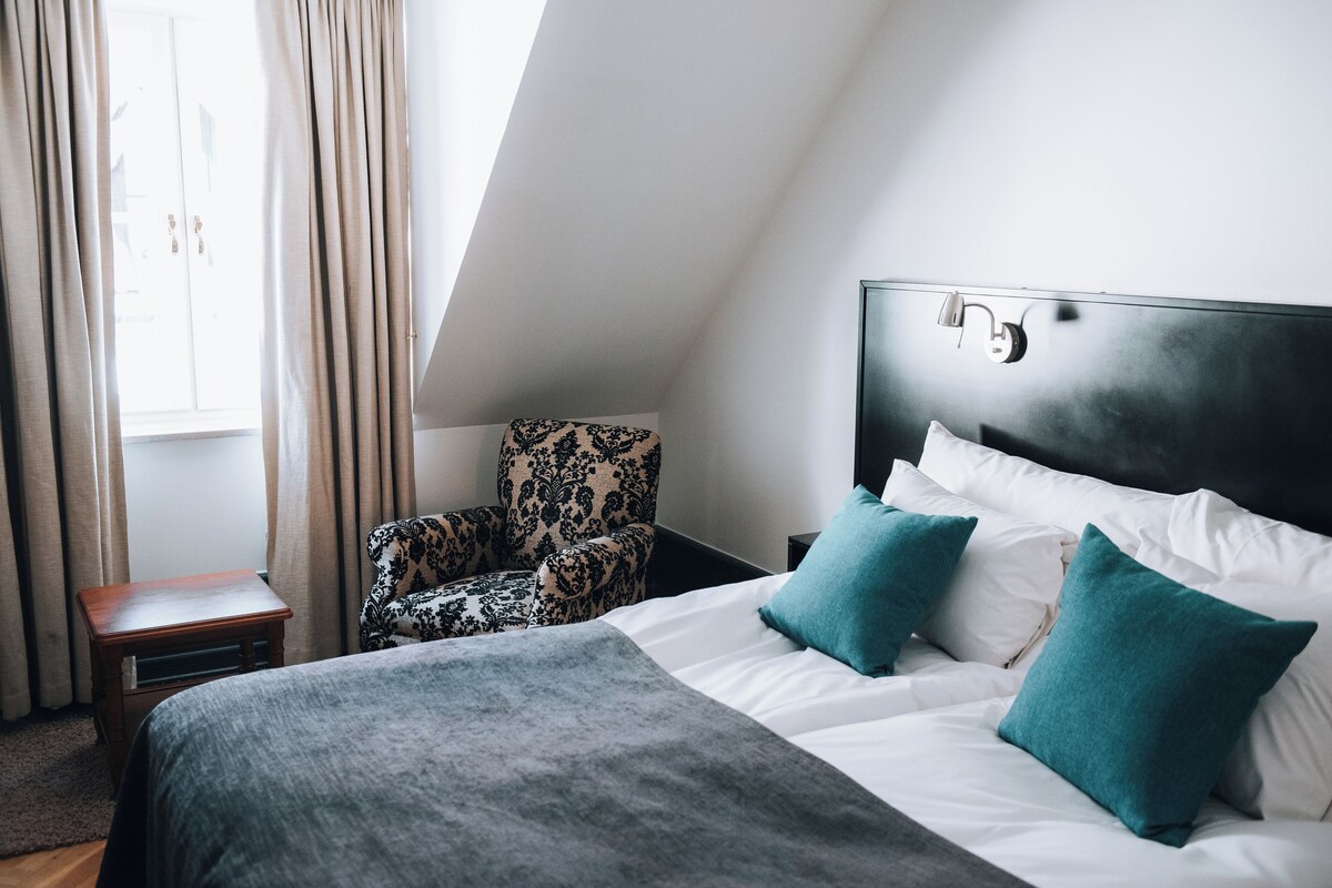 Un dormitorio con cama, una silla, una mesita de noche y una ventana con cortinas.