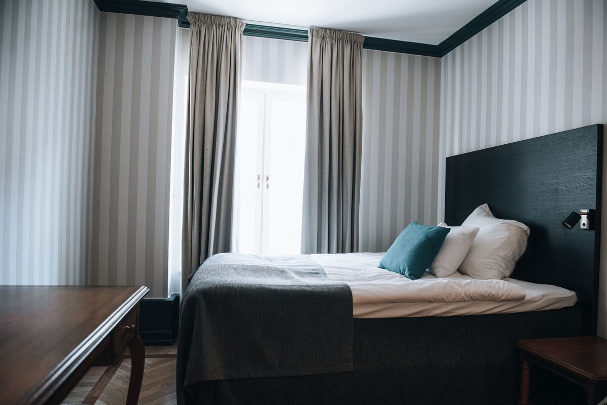 Una habitación de hotel con una cama, una mesita de noche de madera, un escritorio y una ventana con cortinas.