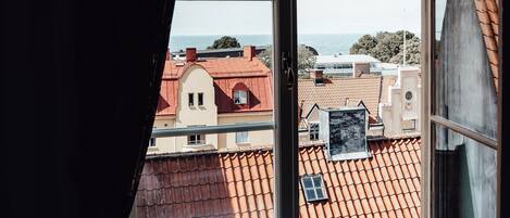 A view from a window with a curtain, overlooking a cityscape with red-roofed buildings and a body of water in the distance.
