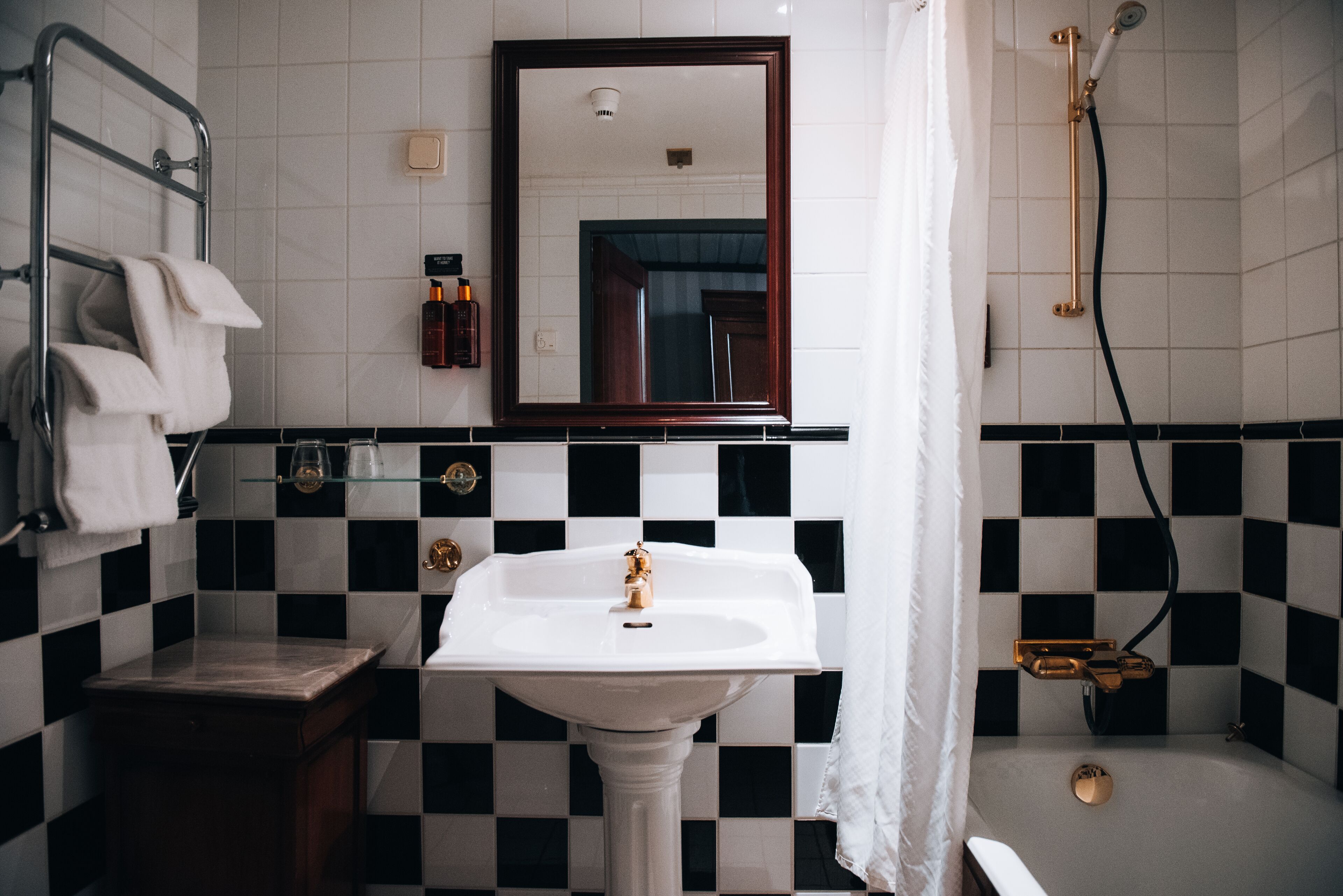 Et badeværelse med væg af skakternet sorte og hvide fliser, en hvid vask med guldfarmet armatur, en glashylde med toiletartikler og en bruseniche med brusehoved i guld.