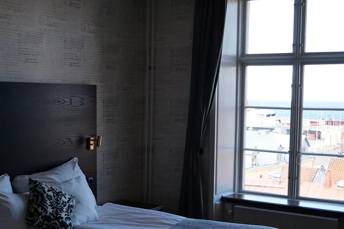 Un dormitorio con una cama grande, un cabecero con texto, una ventana con vistas a edificios y una lámpara colgante.