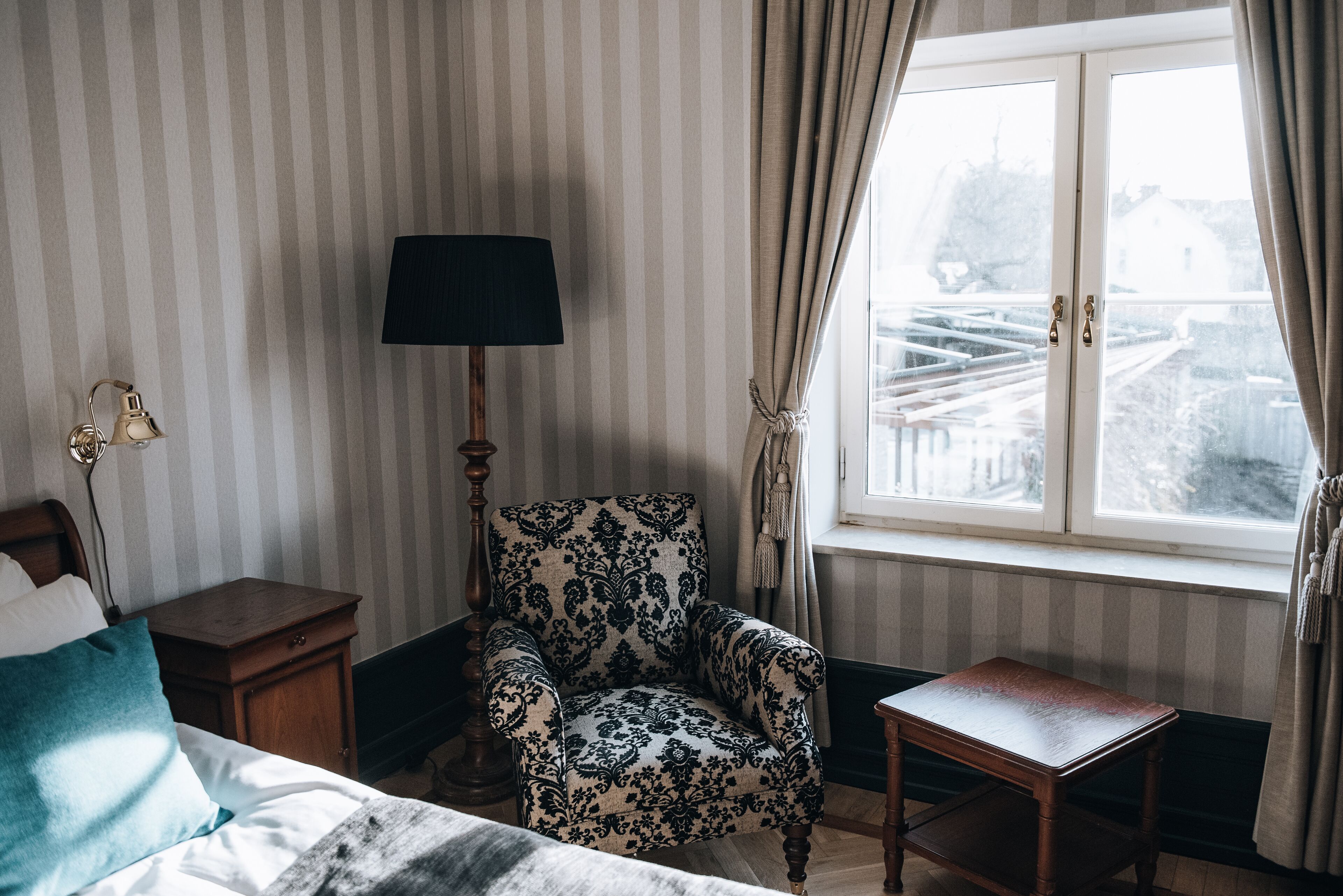 Una habitación con una cama, una silla, una mesita de noche, una lámpara y una ventana con cortinas.