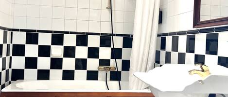 Un baño con una bañera blanca, un inodoro blanco y un lavamanos con grifo dorado. Las paredes están cubiertas de baldosas con un estampado de cuadros blancos y negros.