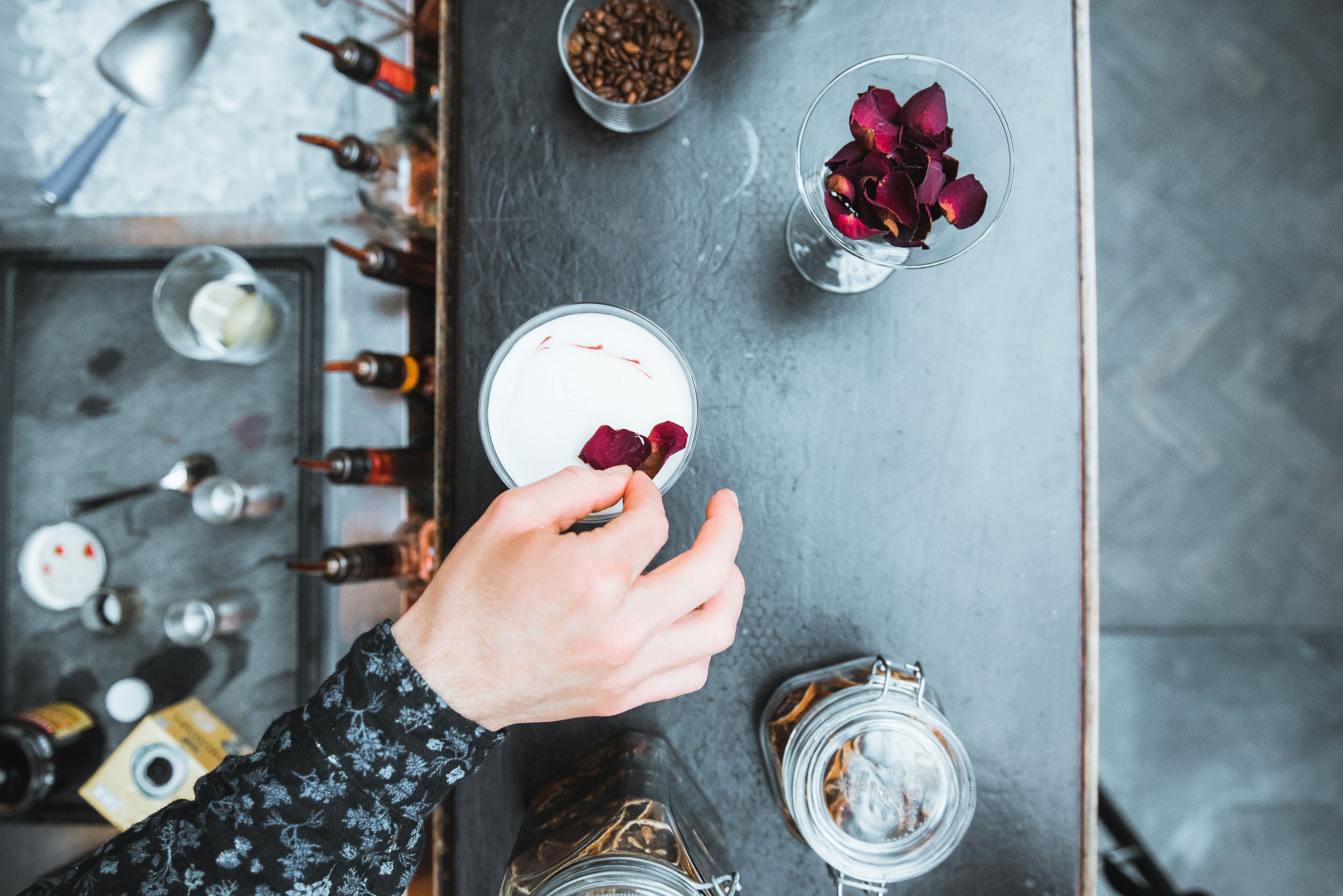 Vue plongeante d’un bar, où l’on aperçoit une main tenant un verre contenant une boisson blanche, ornée d’une fleur rouge, entourée de diverses bouteilles et d’un petit bol de grains de café.