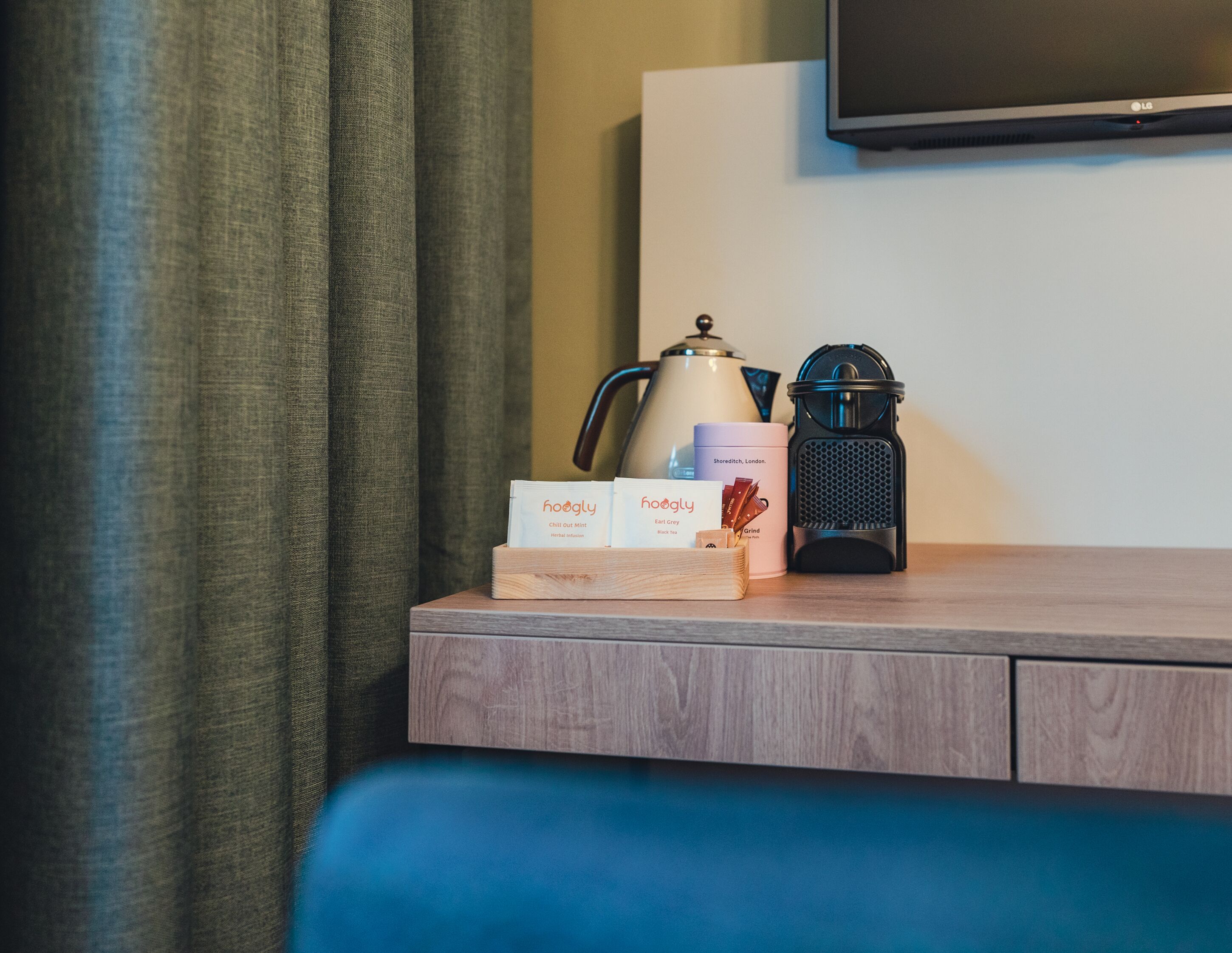A wooden bedside table with a tea kettle, a portable coffee maker, and a box of tea bags.