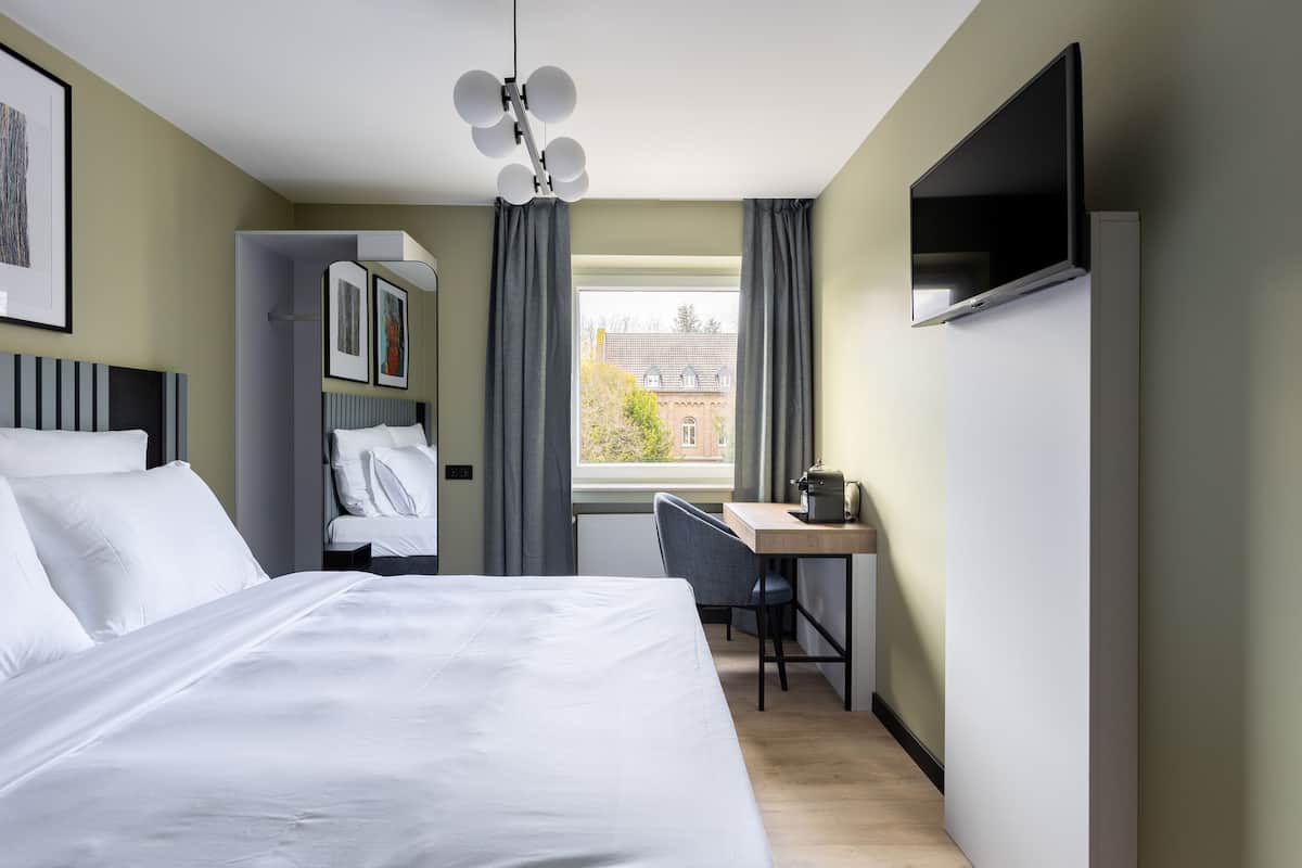 Quarto de hotel com cama, escrivaninha, cadeira, televisão e janela com cortinas.