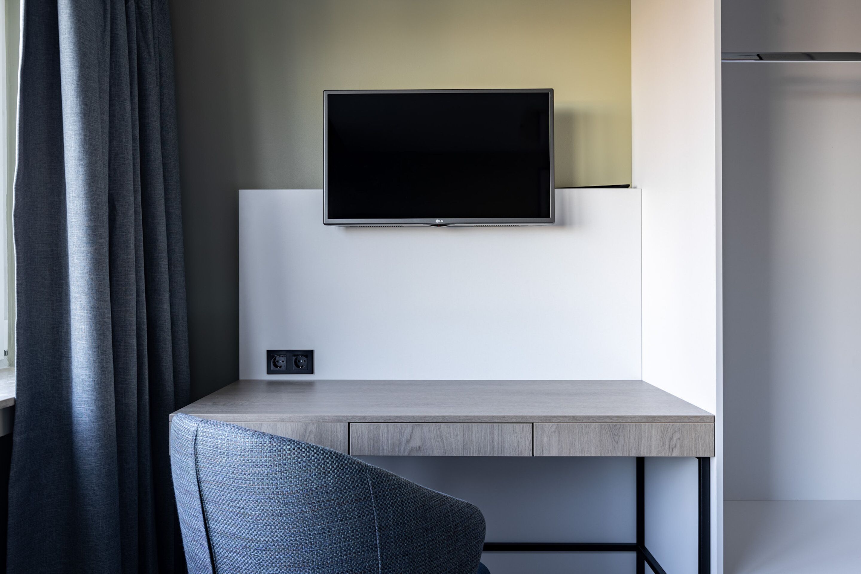 Une chambre moderne avec un téléviseur à écran plat fixé au mur, un bureau de couleur claire et une chaise bleue.