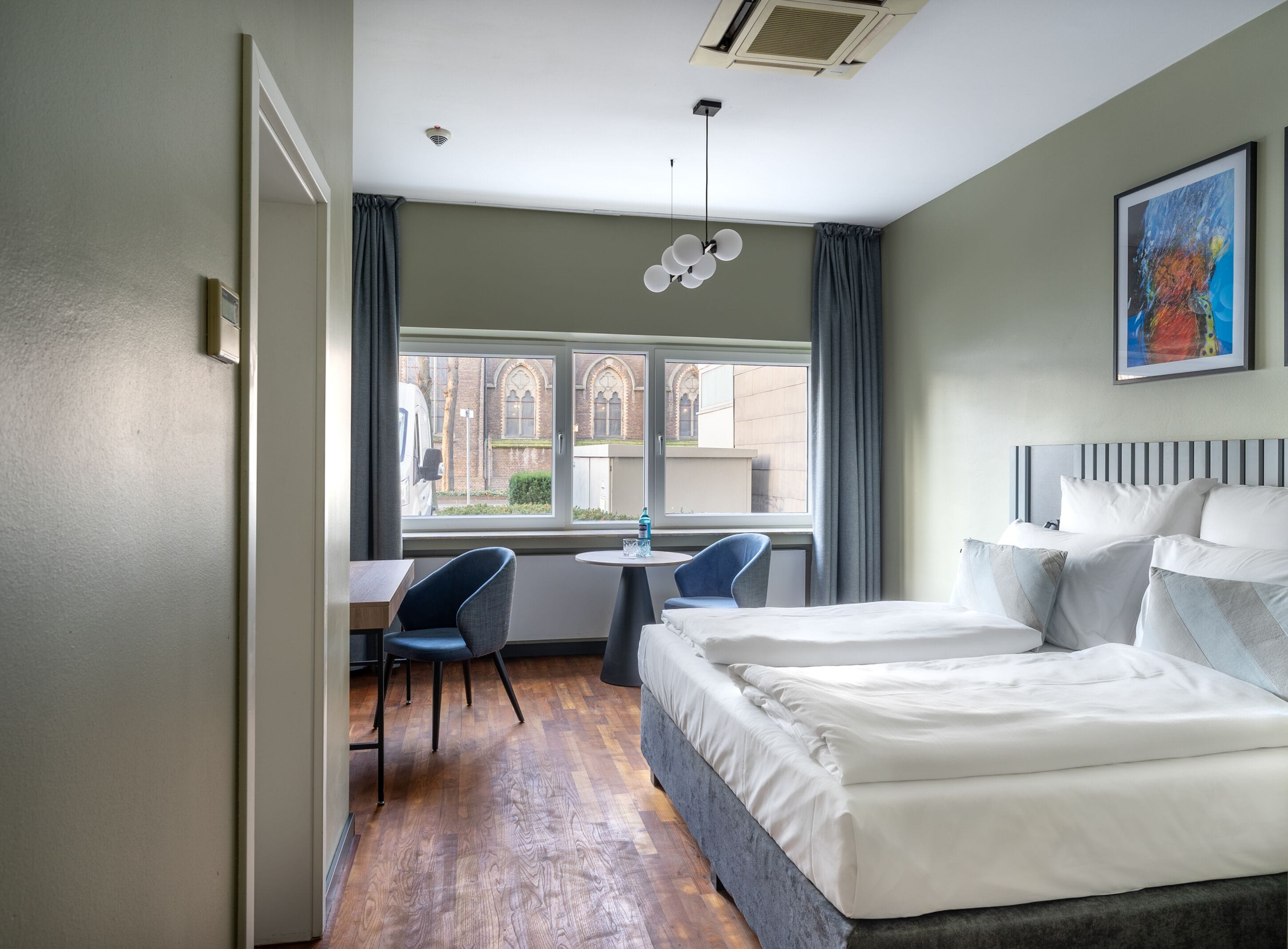 A hotel room with a bed, a desk, two chairs, a window with a view of buildings, and a ceiling fan.