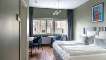 A hotel room with a bed, a desk, two chairs, a window with a view of buildings, and a ceiling fan.