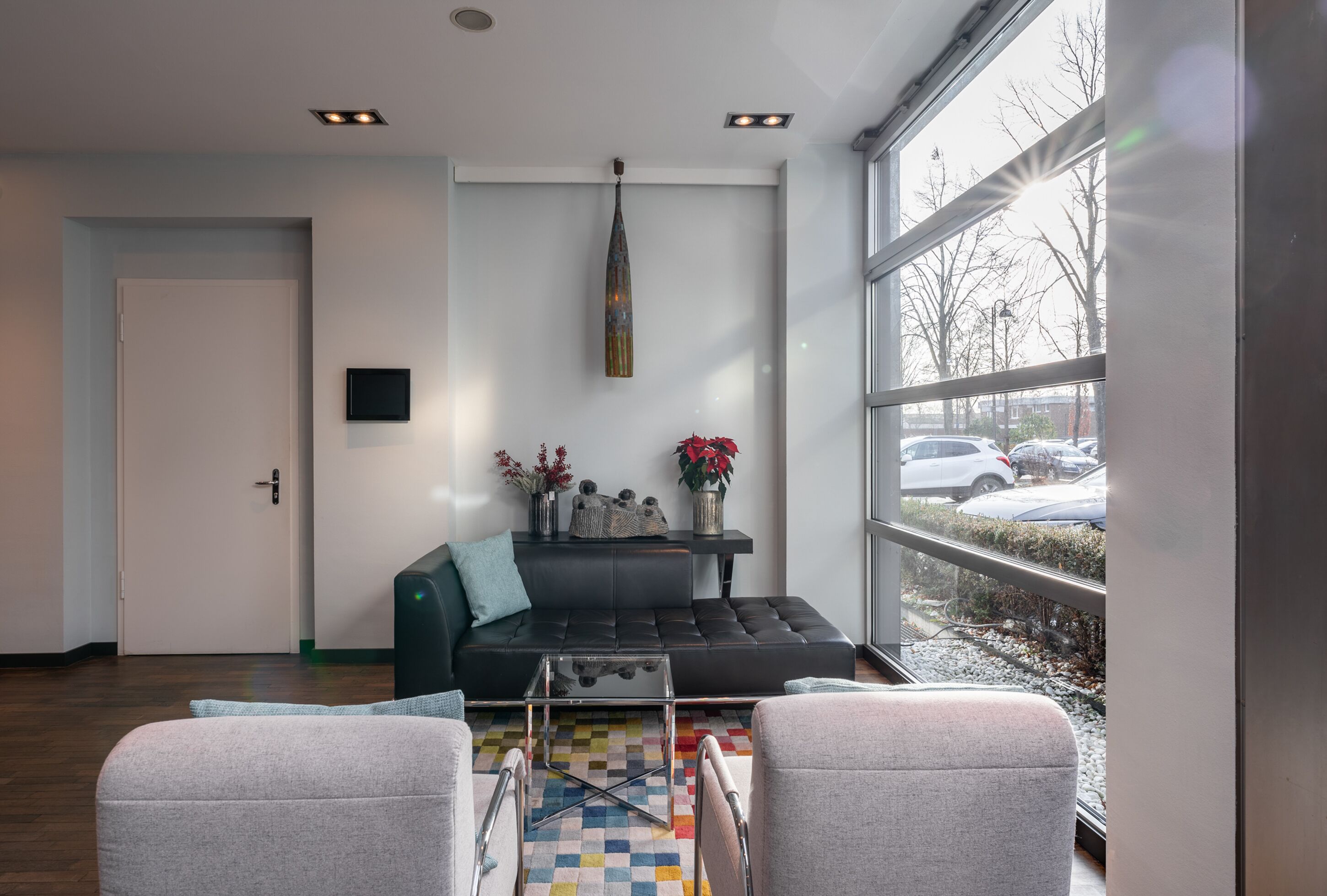 Una sala moderna con un sofá de cuero negro, una mesa de centro de vidrio y dos sillones grises. Se ve un ventanal con vistas a coches estacionados y árboles.