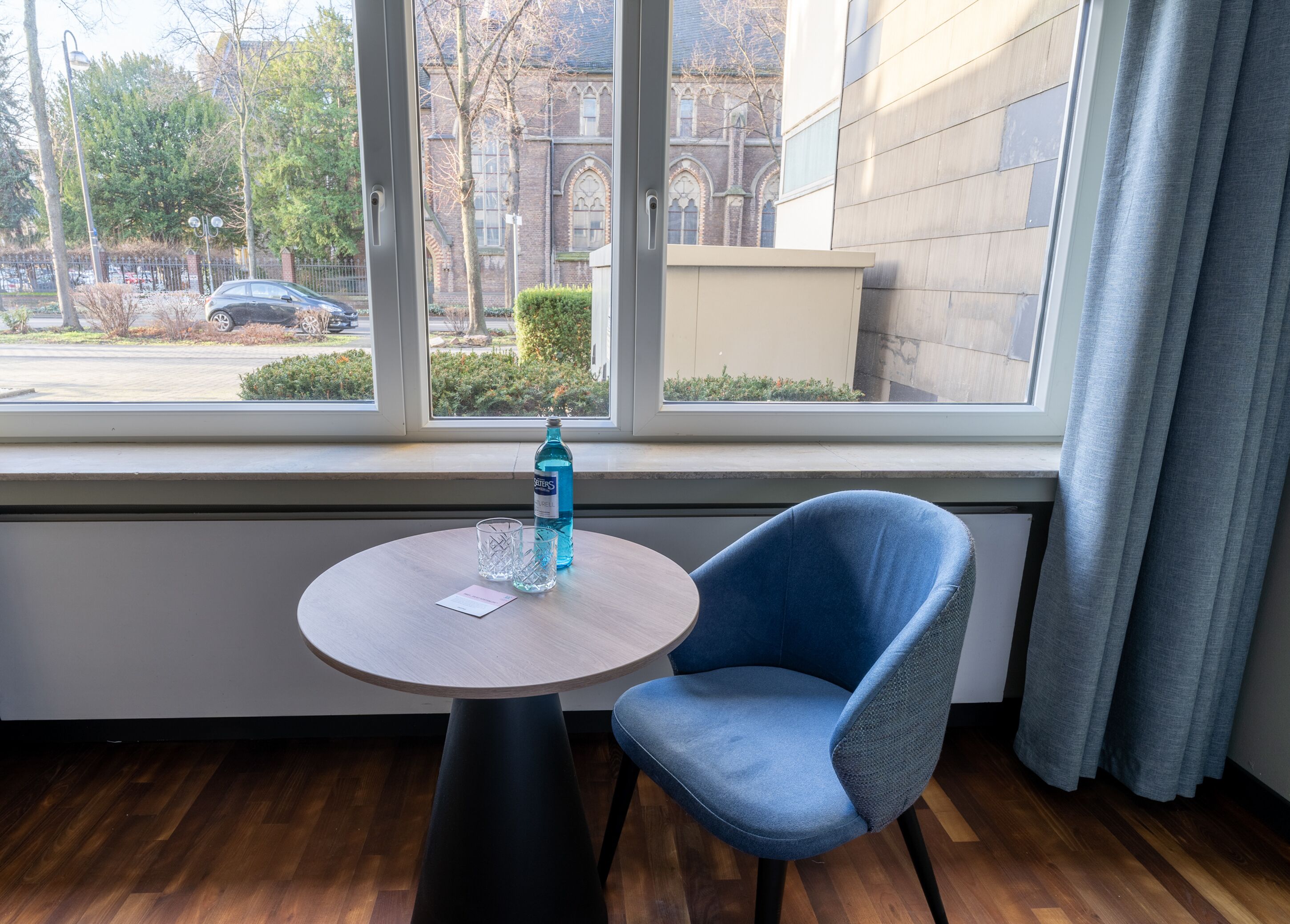 Une pièce avec une table ronde, une chaise bleue, une bouteille d’eau et une fenêtre donnant sur un bâtiment et des arbres.