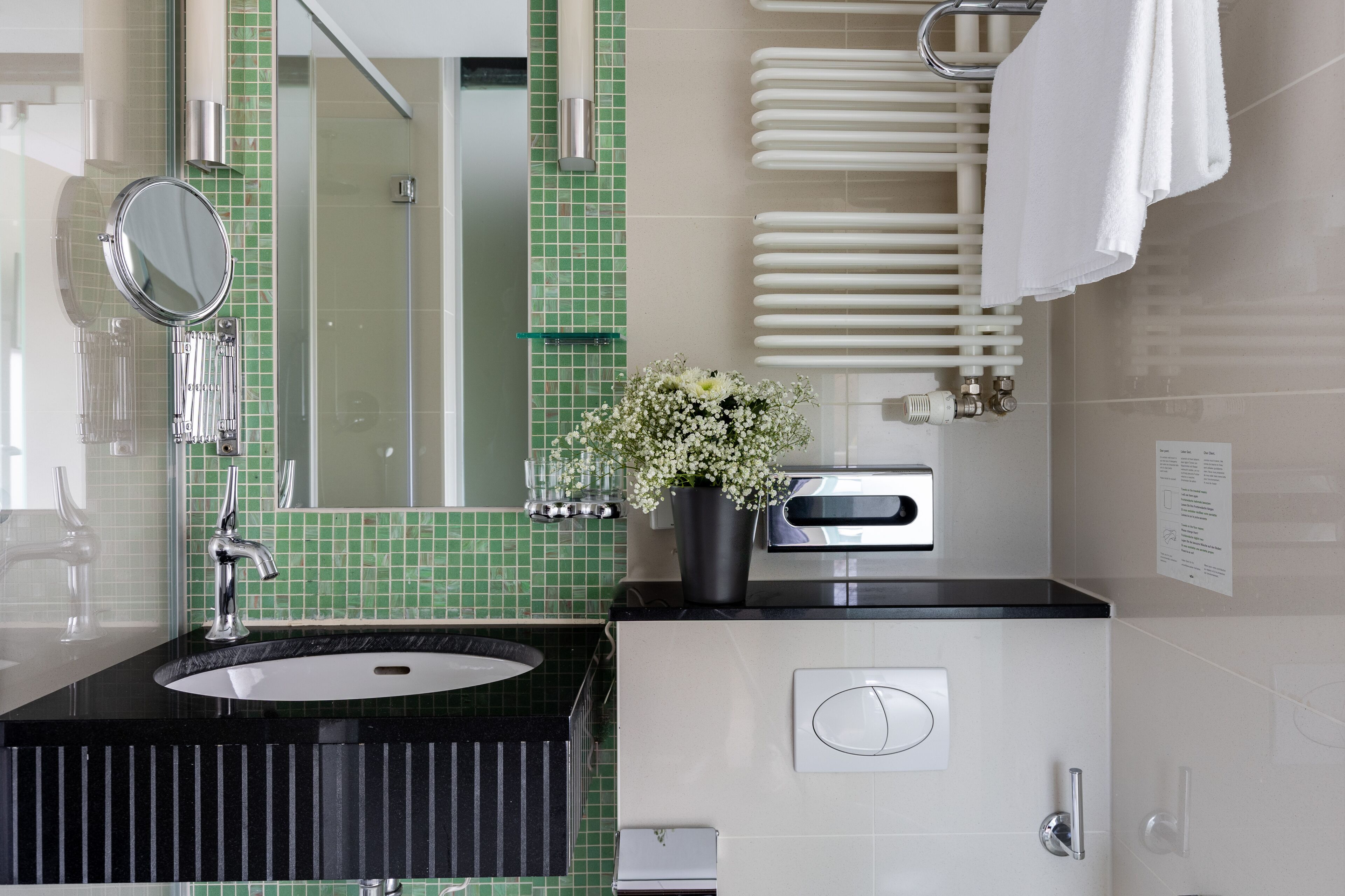 A modern bathroom with a black sink, a mirror, a green tiled backsplash, a white towel rack, and a potted plant.