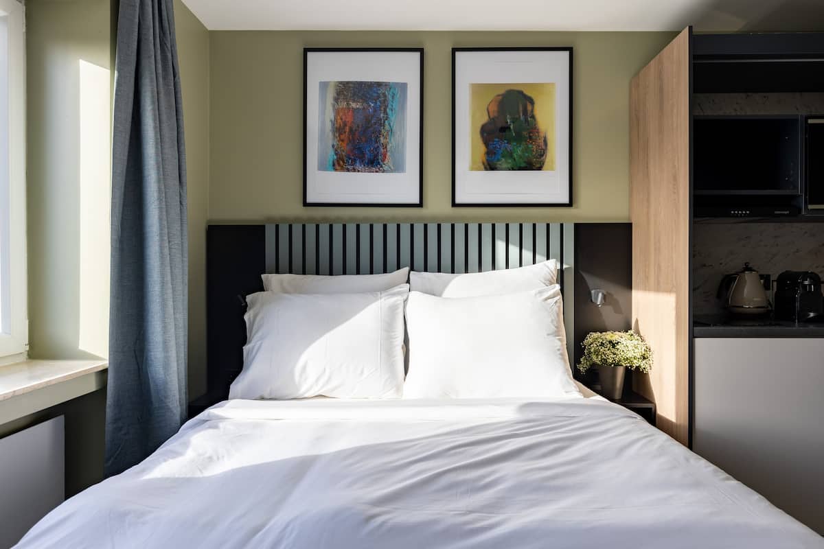 Quarto moderno com cama, duas obras de arte emolduradas, janela com cortinas e área de cozinha.