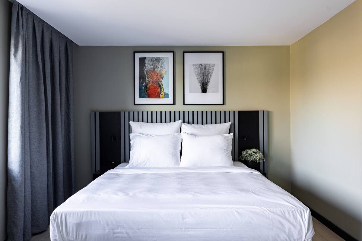 Quarto de hotel com uma cama grande, cortinas cinzentas, duas obras de arte emolduradas na parede e um pequeno vaso com flores.