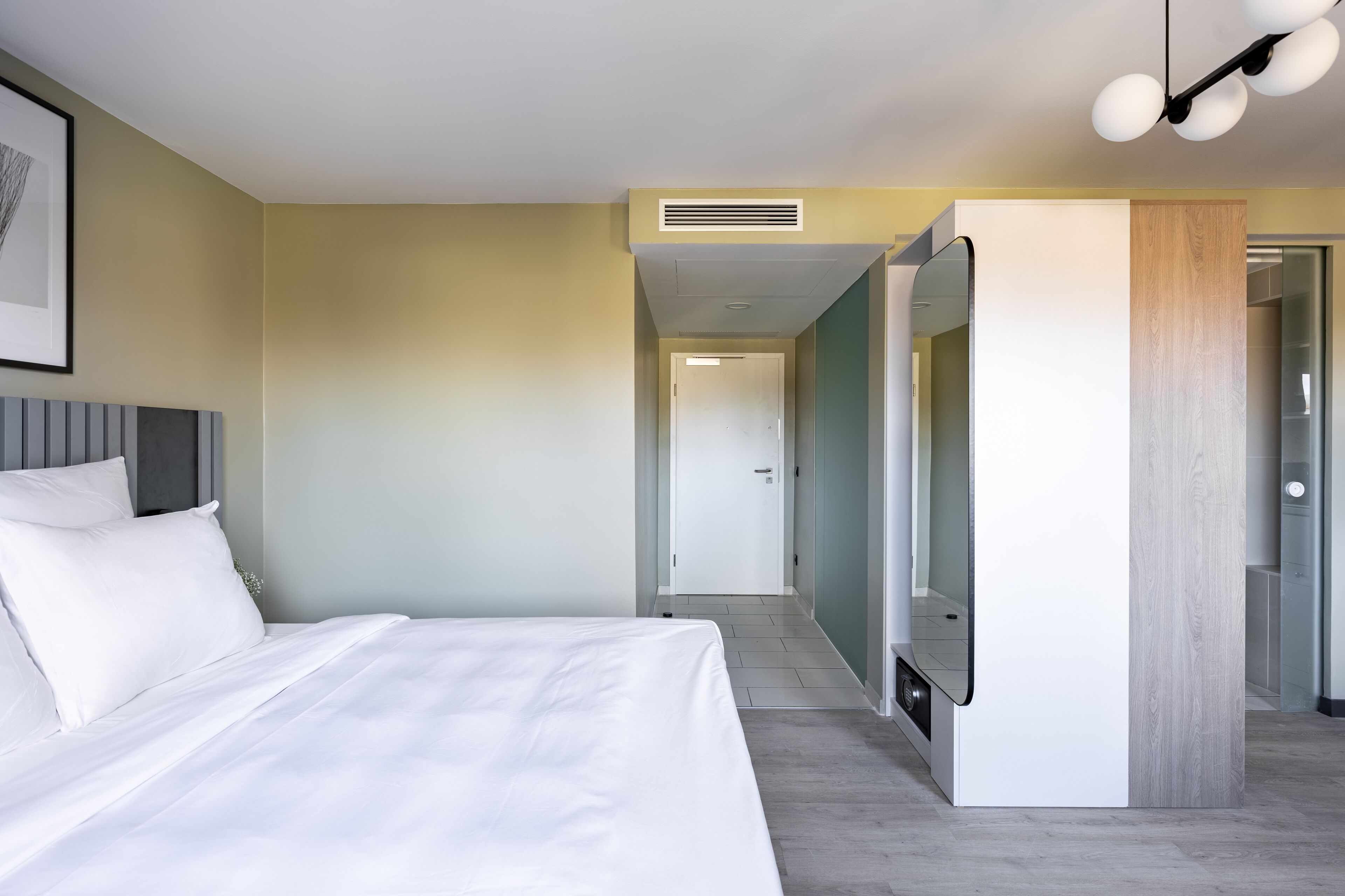 A modern hotel room with a large bed, a flat-screen TV, a wooden accent wall, and a hallway leading to a door.