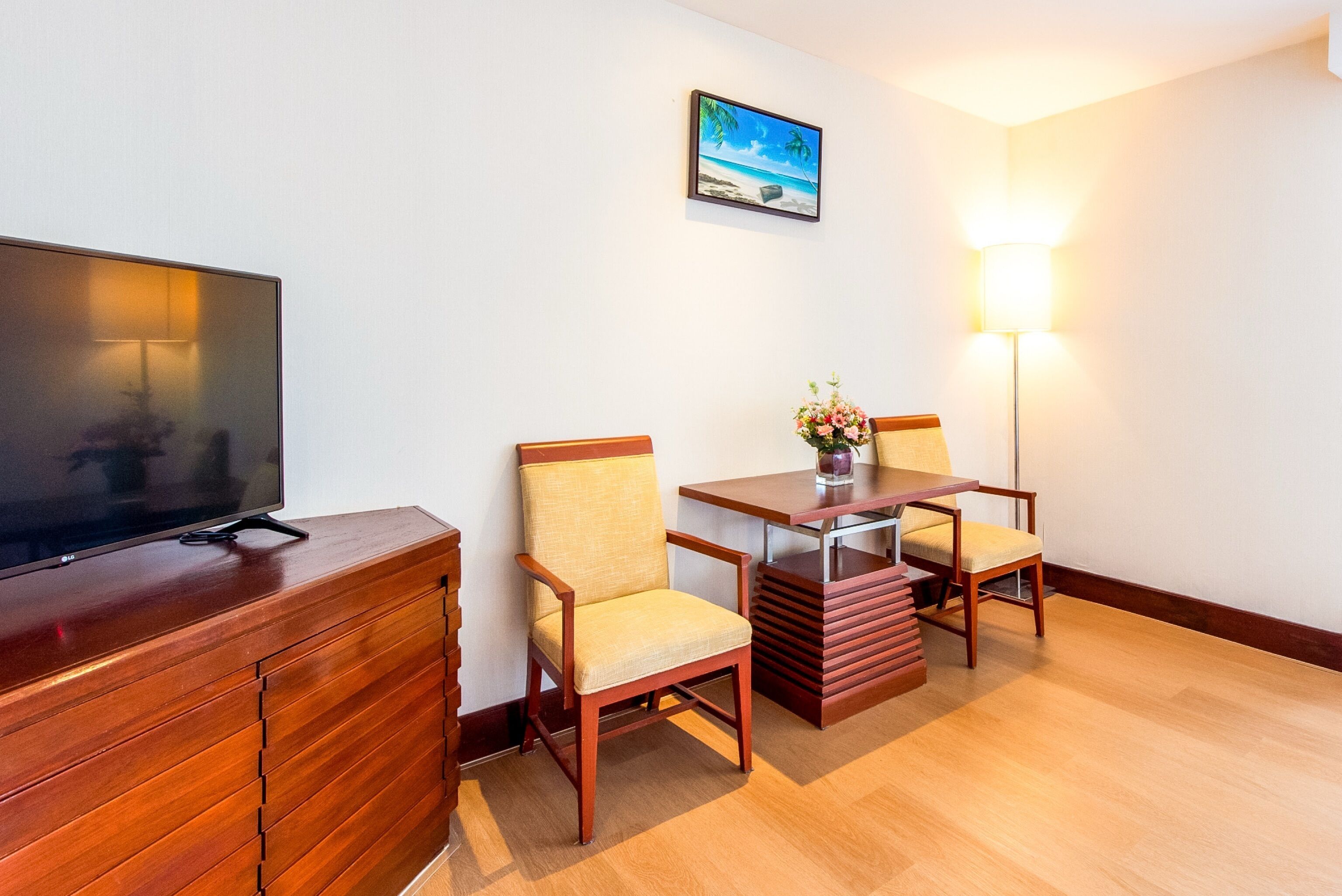 A hotel room with a wooden TV stand, two wooden chairs, a small table with a vase, and a painting on the wall.