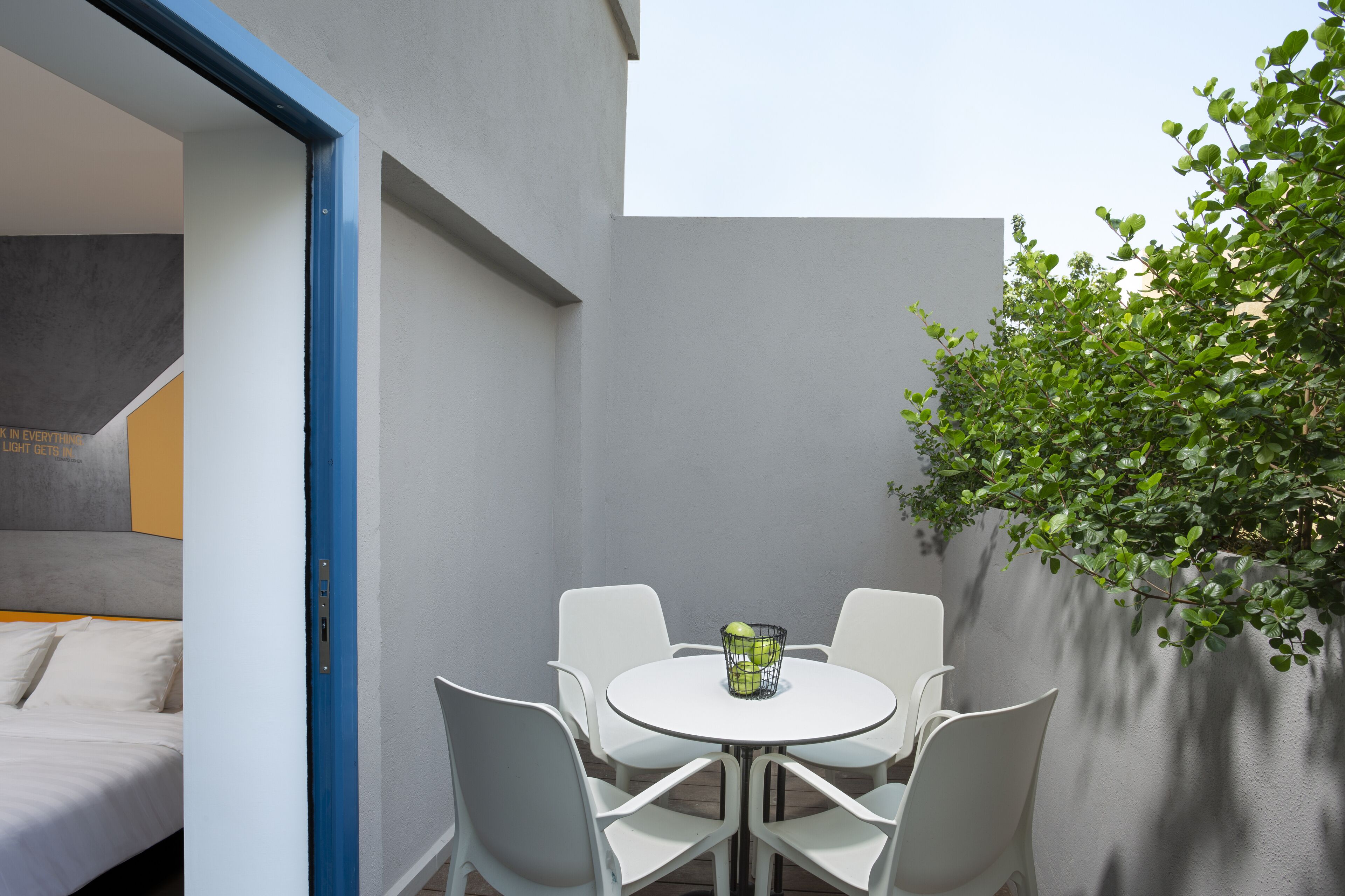 Un espace extérieur moderne, aménagé avec une table ronde et des chaises blanches, une petite plante et un mur orné d’une bordure bleue.