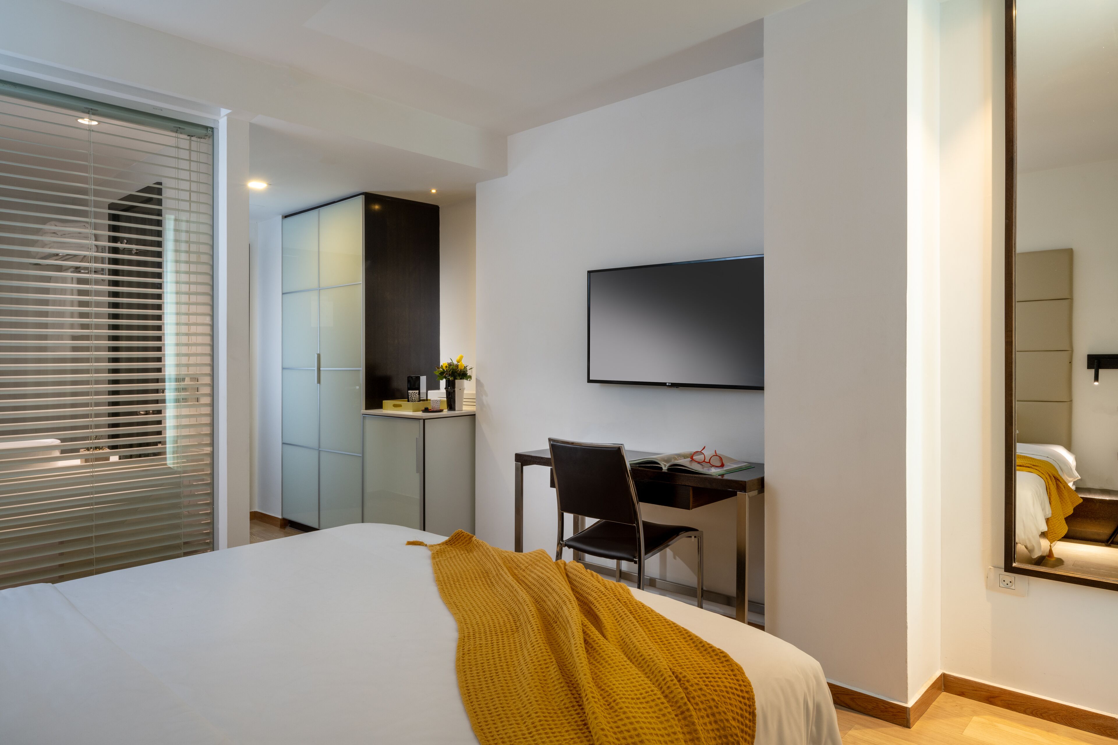 A modern hotel room with a large bed, a desk, a chair, a TV, and a wardrobe.