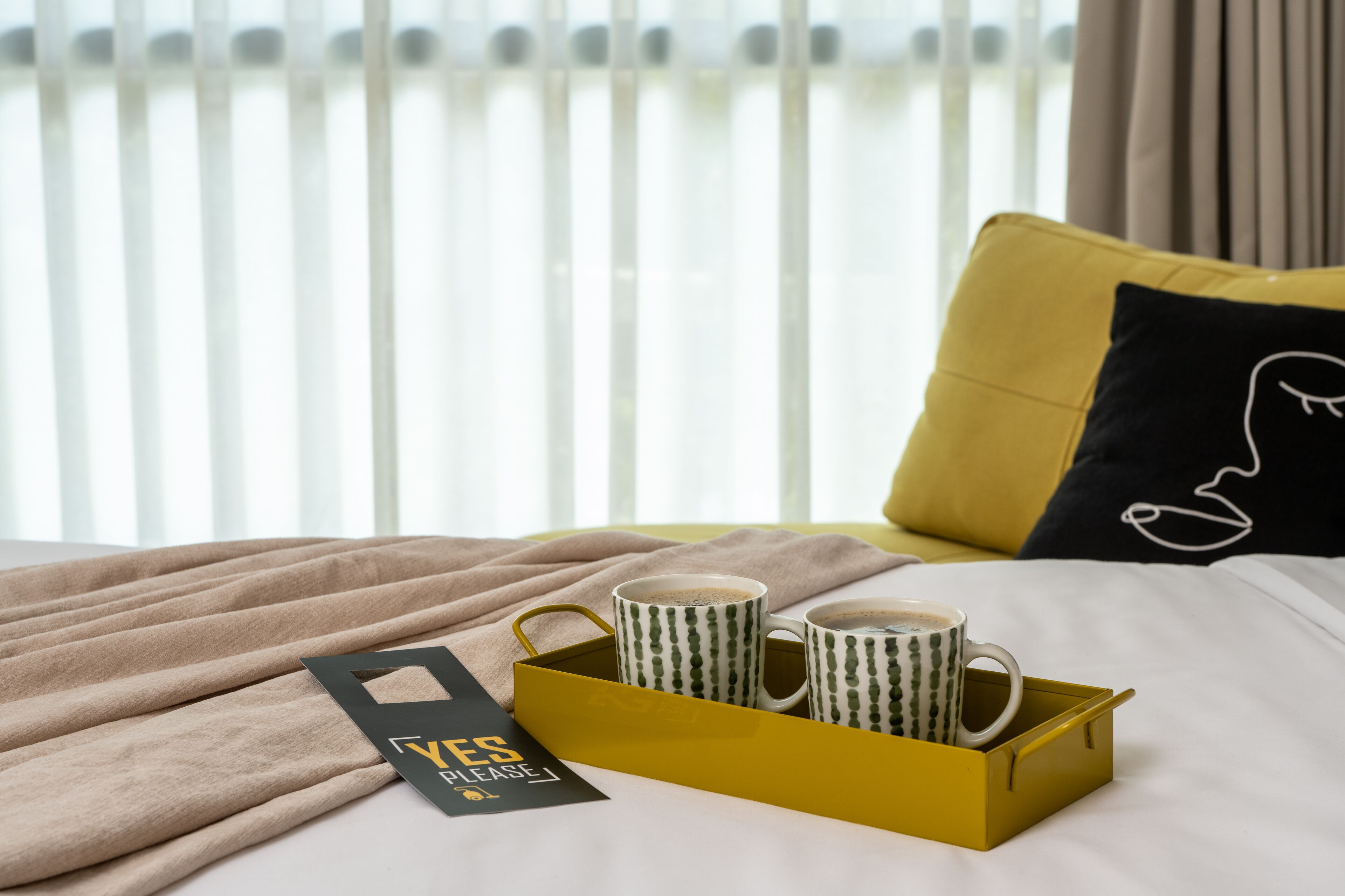 Ein Bett mit einem gelben Tablett, das zwei Tassen enthält, einem schwarzen Kissen mit einem weißen Muster und einem Schild mit der Aufschrift 'YES PLEASE'.