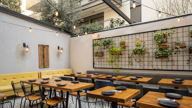 Un restaurante con mesas de madera y sillas de malla negra, un sofá amarillo y una pared decorada con plantas colgantes y un reloj.