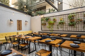 A restaurant with wooden tables and black mesh chairs, a yellow sofa, and a wall with hanging plants and a clock. - BY14 TLV (Tel Aviv)