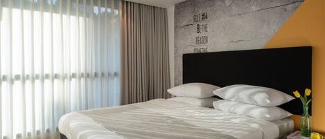 Un dormitorio de hotel moderno con una cama grande, ropa de cama blanca, cabecero negro, mesita de noche con un florero y una frase motivacional en la pared.