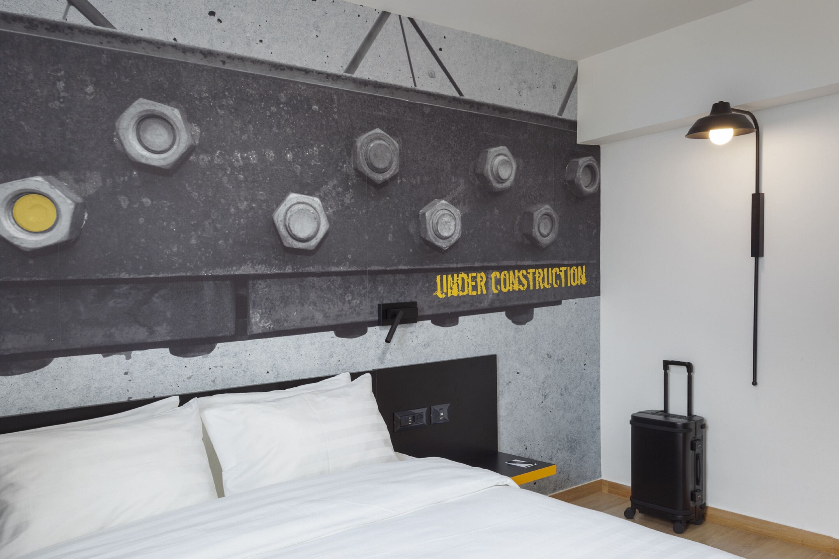 A hotel room with a bed, a headboard with bolts, a wall-mounted lamp, and a suitcase.