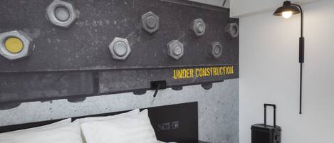 A hotel room with a bed, a headboard with bolts, a wall-mounted lamp, and a suitcase.