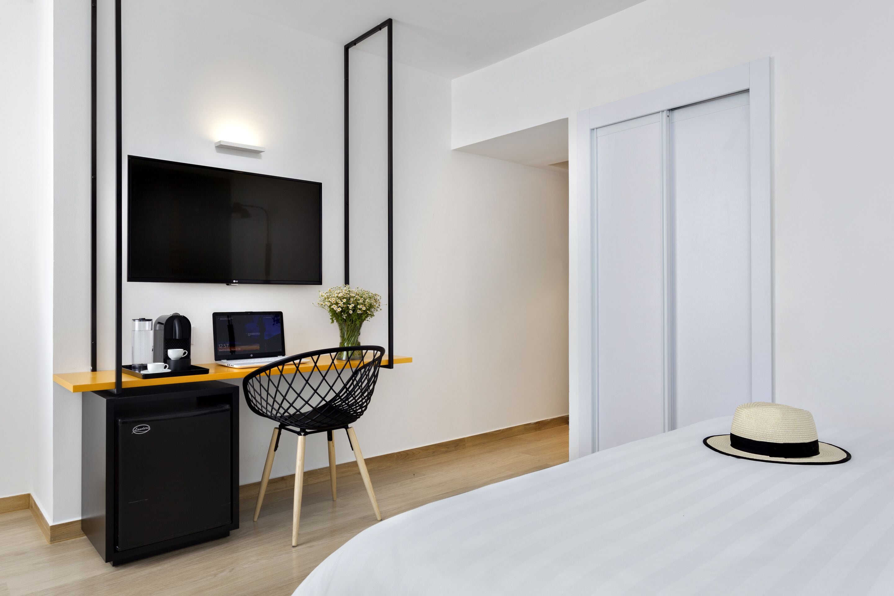 Una habitación de hotel moderna con una cama, un escritorio con computadora, un cafetera, un refrigerador pequeño, un televisor y un florero decorativo.