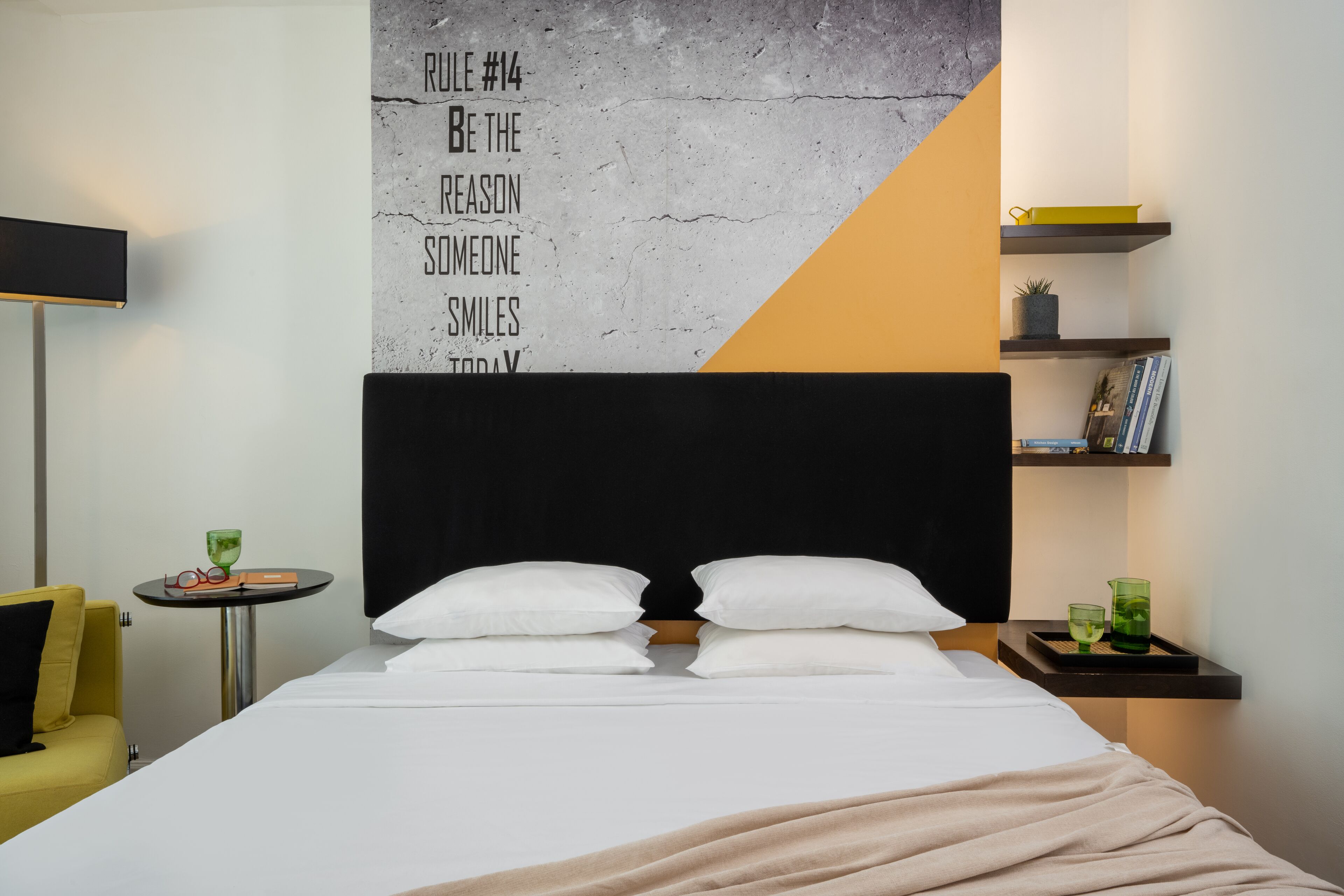 Et hotelværelse med en stor seng, et natbord med en lampe, et lille bord med glas og en bog, samt en væg med motiverende tekst.