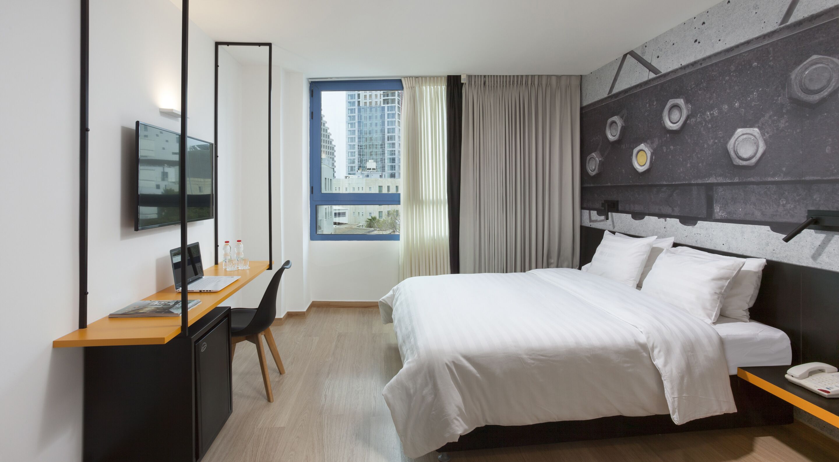 A modern hotel room with a bed, a desk with a laptop, a chair, a TV, and a window with curtains.