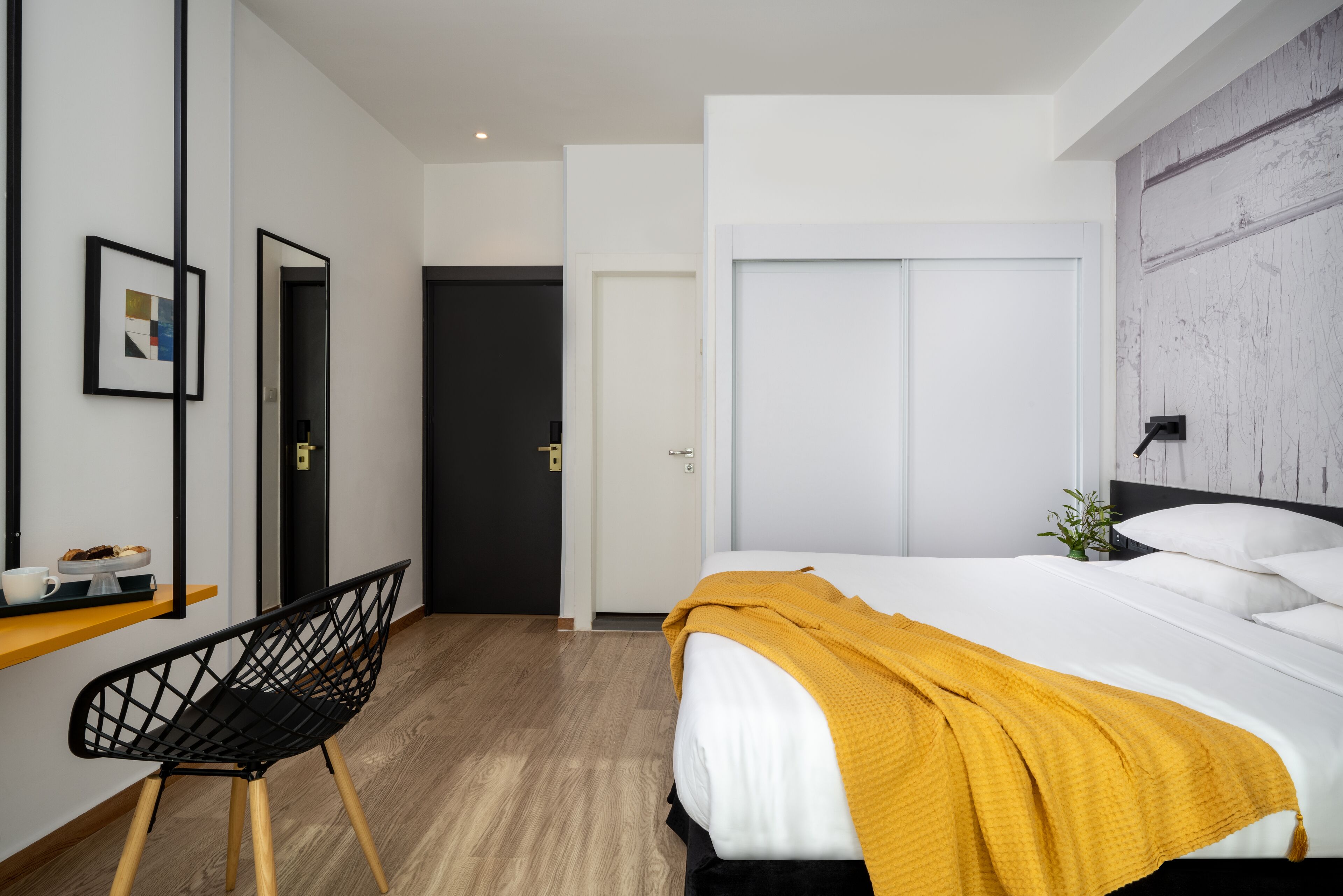 A modern hotel room with a bed, a desk with a chair, a mirror, and a wardrobe.