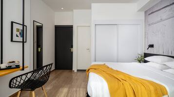 A modern hotel room with a bed, a desk with a chair, a mirror, and a wardrobe.