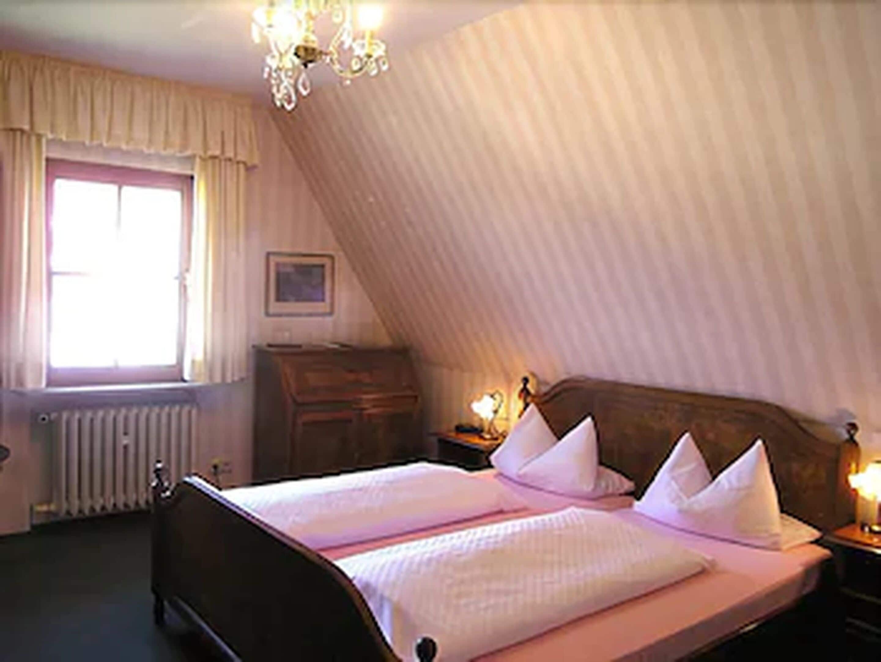 Une chambre à coucher avec un grand lit, une commode, une table de chevet, un lustre, une fenêtre avec des rideaux et un radiateur.