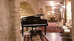 Un piano à queue se trouve dans une pièce spacieuse, dotée d’un plafond haut et de murs en pierre. On y aperçoit des fauteuils en osier et une petite table sur laquelle est posé un vase de fleurs.