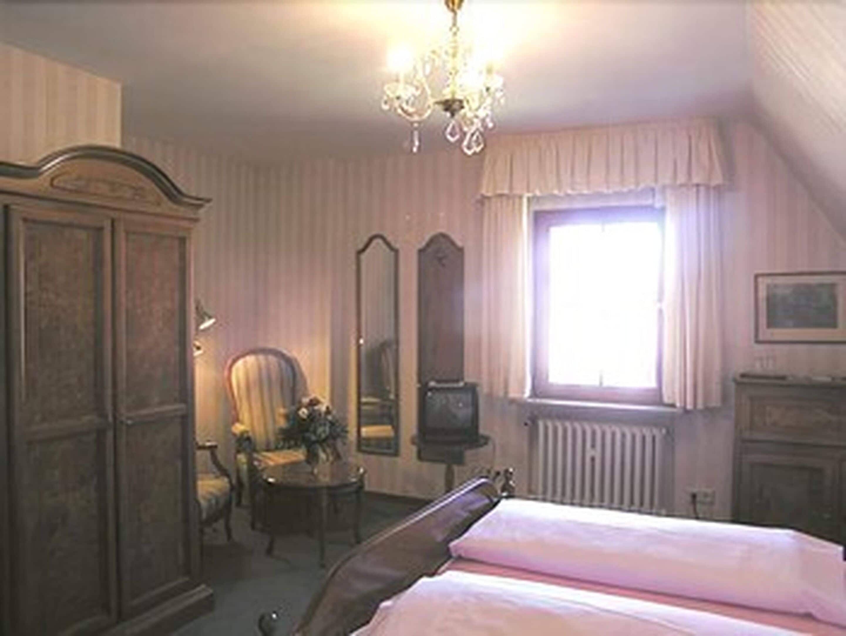 Un dormitorio con cama, una silla, tocador, espejo y ventana con cortinas.