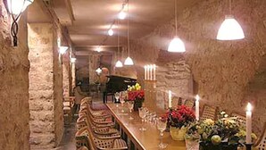 Une longue table à manger, entourée de chaises, agrémentée de bougies et de compositions florales, dans une pièce rustique aux murs de pierre, illuminée par des lampes suspendues.