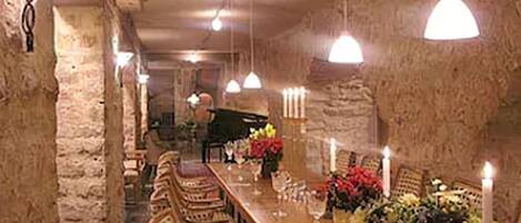Una larga mesa de comedor con sillas, velas y centros de mesa florales en una sala rústica de paredes de piedra con luces colgantes.