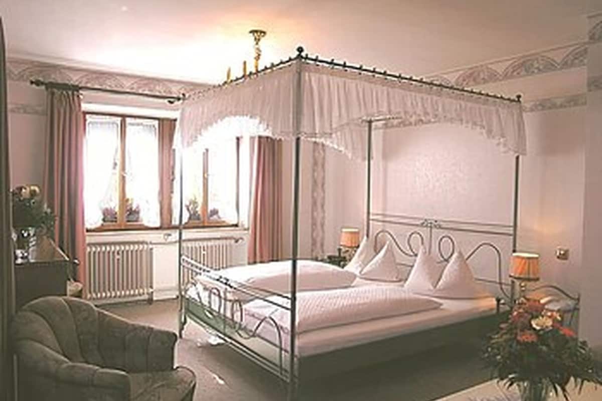 A four-poster canopy bed with white linens, a bedside table with a lamp, and a chair in a room with a radiator and curtains.